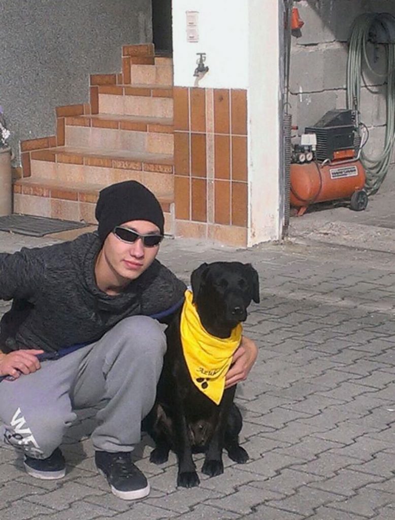Marco trägt Mütze und Sonnenbrille. Er kniet mit seinem neuen, schwarzen Blindenhund in einer Einfahrt