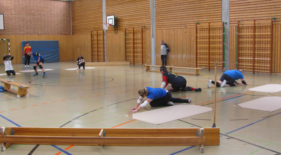 Im Vordergrund kniet das verteidigende Team vor ihren Matten. Im Hintergrund macht sich ein Angreifer bereit für den Wurf