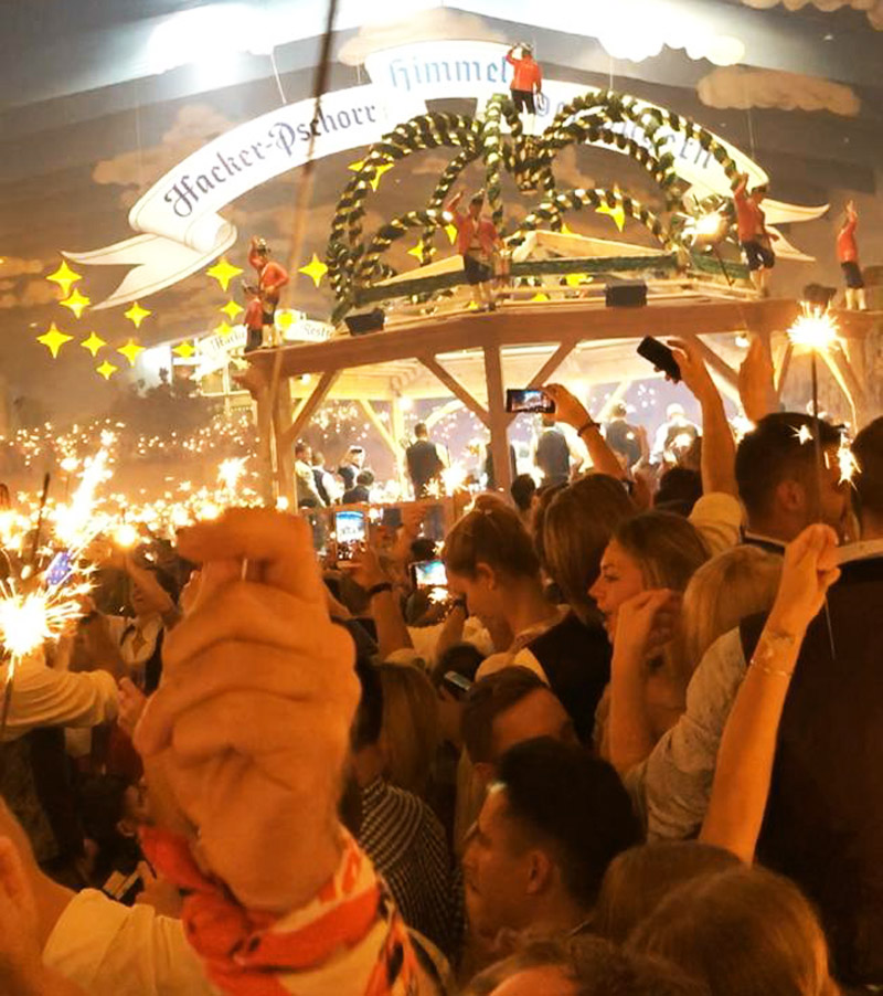 Menschenmenge im Hackerzelt auf dem Oktoberfest, wunderkerzen brennen in vielen Händen, über den Köpfen der Leute leuchtet der Logobanner "Hacker-Pschorr-Himmel