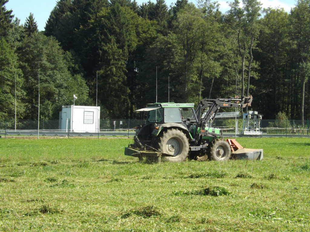 Ein Traktor mit Kreiselmäher fährt auf einer Wiese vor einer eingezäunten Meßstation für Klimaforschung im oberbayerischer Weiler Fendt