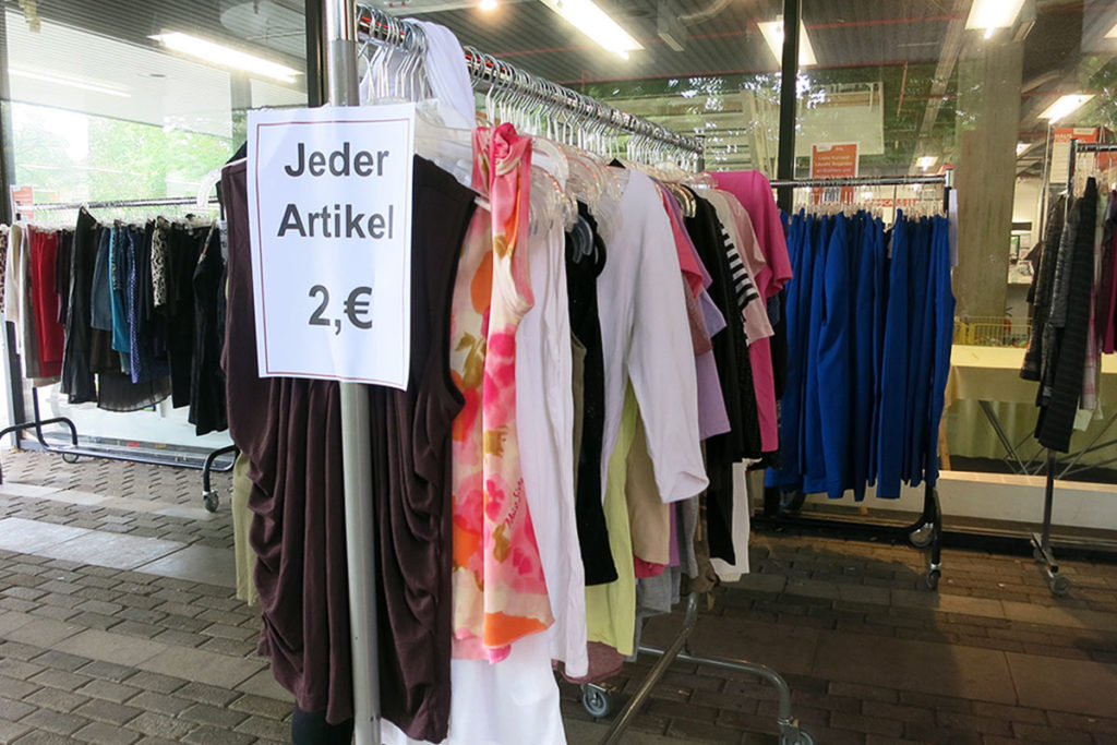 Kleiderstangen, vollgehängt mit Kleidern, Hosen, Röcken stehen vor der diakonia. Auf dem großen Preisschild steht: Jeder Artikel 2, €