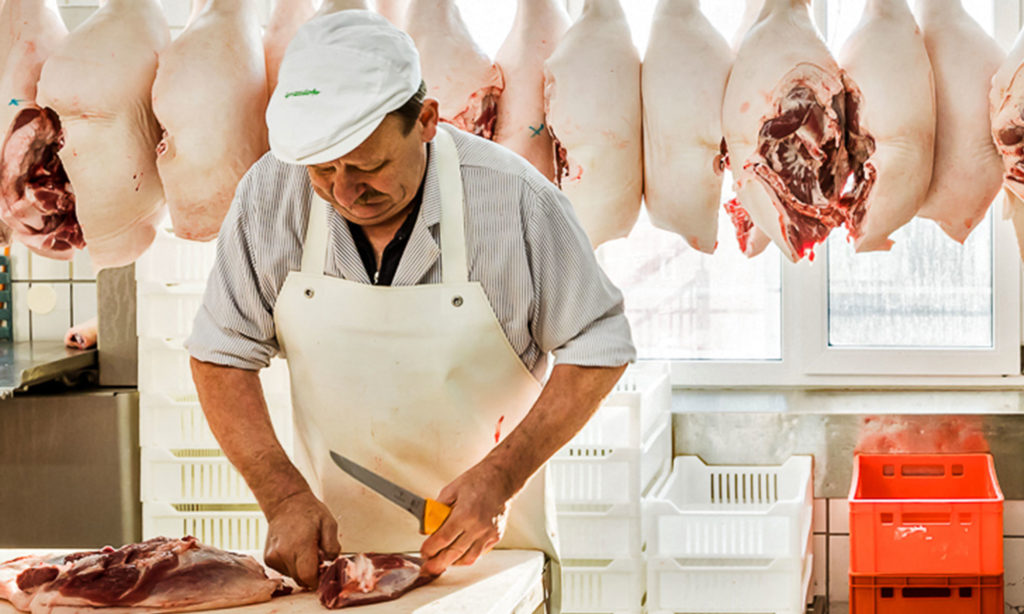 Ein Metzger zerlegt Fleisch in der hofeigenen Metzgerei. Im Hintergrund hängen Fleischhälften.