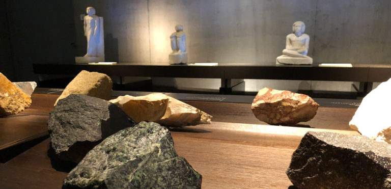 Blick in Raum "Ägypten (er)fassen" im Ägyptischen Museum München. Angeschnittene Steine im Vordergrund. Hinten beleuchtete Skulpturen. Selbsttest "Blind Kunst sehen" von Tanja Praske für Freiraum