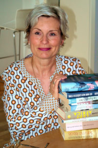 Franziska Feind sitzt vor einem Bücherstapel an einem Tisch. Sie trägt eine weiße Bluse mit braunen Punkten und lächelt in in die Kamera. Foto: Nina Jarosch