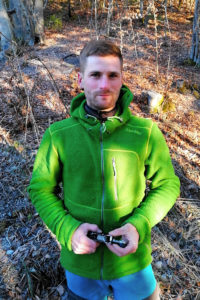 Jan-Marco Donat ist Baumpfeger. Er trägt eine grüne Fleece-Jacke und blaue Arbeitshosen und steht in einem Wald. In der Hand hält er einen Karabinerhaken, den er zum Klettern benötigt. Foto: Nina Jarosch