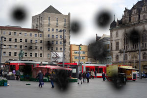 Blick auf einen Marktplatz bei trübem Wetter. Im Vordergrund laufen einige Menschen zwischen Verkaufsständen. Im Hintergrund fährt eine Straßenbahn vor einem Gebäude entlang. Das Bild ist durch vereinzelte Flecken verdeckt. Foto: Jonas Kessel