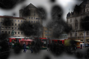 Blick auf einen Marktplatz bei trübem Wetter. Im Vordergrund laufen einige Menschen zwischen Verkaufsständen. Im Hintergrund fährt eine Straßenbahn vor einem Gebäude entlang. Das Bild ist durch zahlreiche Flecken verdeckt und fast nicht mehr zu erkennen. Foto: Jonas Kessel