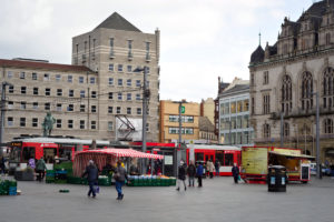 Blick auf einen Marktplatz bei trübem Wetter. Im Vordergrund laufen einige Menschen zwischen Verkaufsständen. Im Hintergrund fährt eine Straßenbahn vor einem Gebäude entlang. Foto: Jonas Kessel