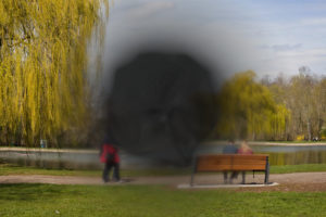 Blick auf einen Park mit Teich und Wasserfontäne. Im Vordergrund sitzt ein junges Pärchen auf einer Parkbank. Von links läuft ein älteres Pärchen durch das Bild. In Vorder- und Hintergrund sind Weidenbäume zu sehen. Der Himmel ist bewölkt, aber die Sonne scheint. Das zentrale Gesichtsfeld ist verzerrt, verdunkelt und nicht mehr zu erkennen. Foto: Jonas Kessel