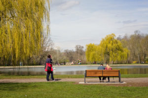 Blick auf einen Park mit Teich und Wasserfontäne. Im Vordergrund sitzt ein junges Pärchen auf einer Parkbank. Von links läuft ein älteres Pärchen durch das Bild. In Vorder- und Hintergrund sind Weidenbäume zu sehen. Der Himmel ist bewölkt, aber die Sonne scheint. Foto: Jonas Kessel