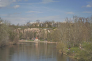 Blick auf einen Fluss mit einer Felsklippe und Häusern im Hintergrund. Der Flusslauf wird von Bäumen flankiert. Der Himmel ist blau und die Sonne scheint. Das Bild ist kontrastarm und etwas verschwommen. Foto: Jonas Kessel