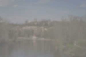 Blick auf einen Fluss mit einer Felsklippe und Häusern im Hintergrund. Der Flusslauf wird von Bäumen flankiert. Der Himmel ist blau und die Sonne scheint. Das Bild hat fast keinen Kontrast mehr und ist stark verschwommen. Foto: Jonas Kessel