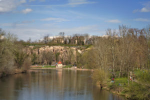 Blick auf einen Fluss mit einer Felsklippe und Häusern im Hintergrund. Der Flusslauf wird von Bäumen flankiert. Der Himmel ist blau und die Sonne scheint. Foto: Jonas Kessel