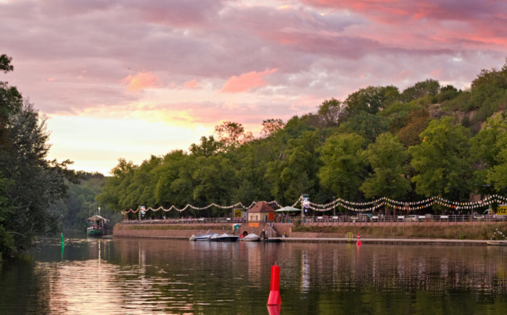 Blick von der Ziegelwiese des Peißnitzparks in Halle auf das Riveufer, ein befestigte Uferanlage aus Sand- und Backstein. Der rosafarbene Sonnenuntergang taucht den Fluss, die Bäume und die dahinterliegende Feldklippe in kitschiges Licht. Am Ufer sind Laternenreihen aufgehangen. Foto: Jonas Kessel