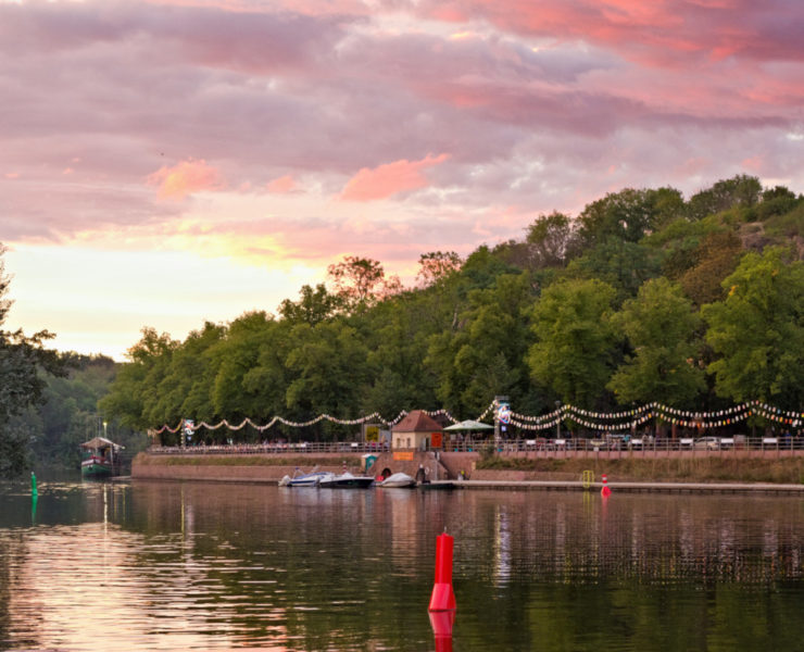 Blick von der Ziegelwiese des Peißnitzparks in Halle auf das Riveufer, ein befestigte Uferanlage aus Sand- und Backstein. Der rosafarbene Sonnenuntergang taucht den Fluss, die Bäume und die dahinterliegende Feldklippe in kitschiges Licht. Am Ufer sind Laternenreihen aufgehangen. Foto: Jonas Kessel