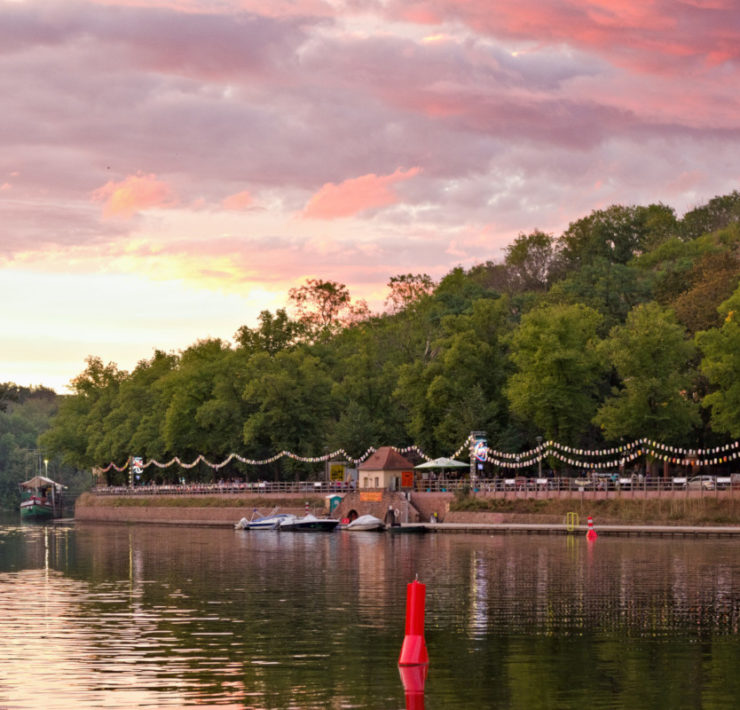 Blick von der Ziegelwiese des Peißnitzparks in Halle auf das Riveufer, ein befestigte Uferanlage aus Sand- und Backstein. Der rosafarbene Sonnenuntergang taucht den Fluss, die Bäume und die dahinterliegende Feldklippe in kitschiges Licht. Am Ufer sind Laternenreihen aufgehangen. Foto: Jonas Kessel