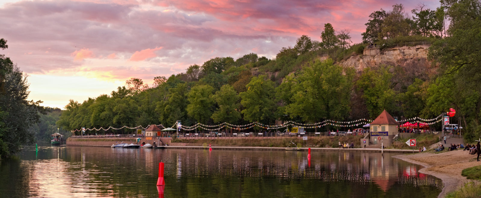 Blick von der Ziegelwiese des Peißnitzparks in Halle auf das Riveufer, ein befestigte Uferanlage aus Sand- und Backstein. Der rosafarbene Sonnenuntergang taucht den Fluss, die Bäume und die dahinterliegende Feldklippe in kitschiges Licht. Am Ufer sind Laternenreihen aufgehangen. Foto: Jonas Kessel