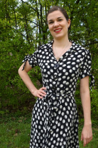 Sandra Brand trägt ein schwarzes Kleid mit weißen Punkten im Vintage-Stil. Sie stützt eine Hand in die taille und lächelt adrett in die Kamera. Sie steht in einem Park. Foto: Nina Jarosch