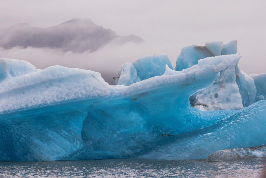 Gletschereis das hellblau leuchtet, im Vordergrund offenes Wasser, im Hintergrund Berge im Nebel