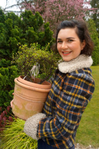 Victoria Stettnersteht im Garten und trägt eine karierte Winterjacke, lächelt in die Kamera und hat einen Blumentopf in den Händen. Foto: Nina Jarosch