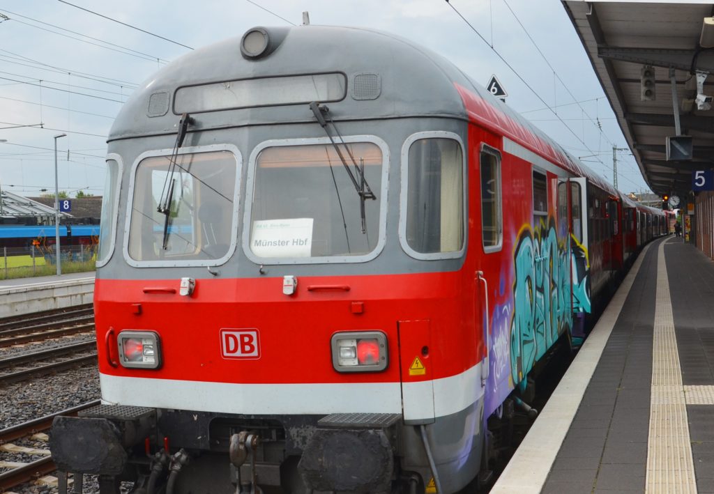 Ein rot-grauer Zug besprüht an der rechten Seite mi Graffiti steht am Gleis 5, im Hintergrund grauer Himmel