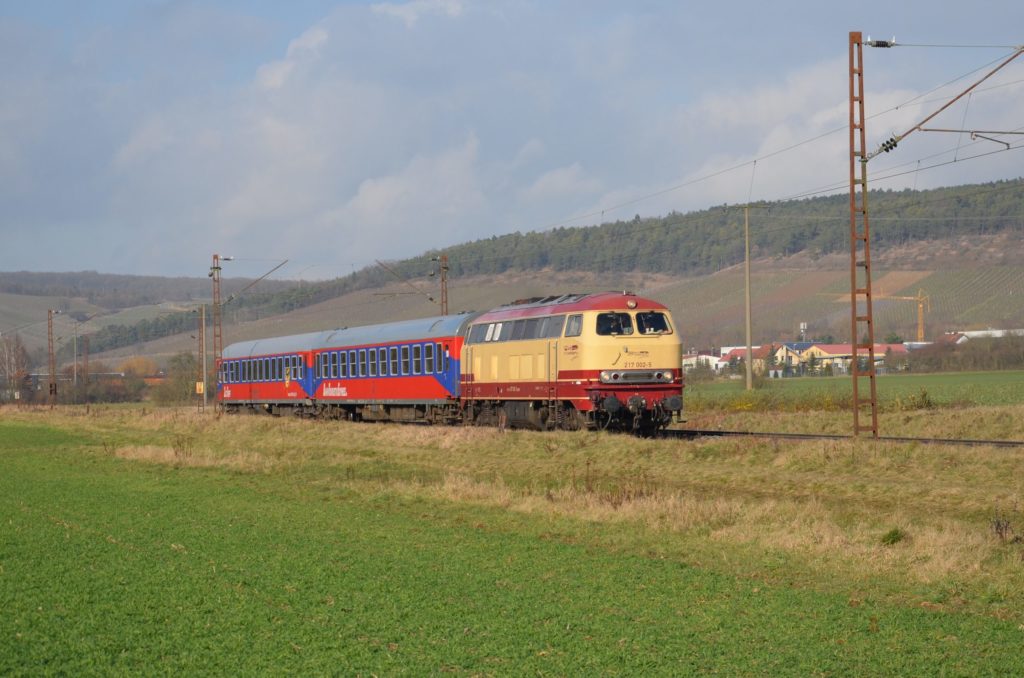 Ein Zug mit einer beige-braunen alten Lok fährt durch eine grüne Landschaft