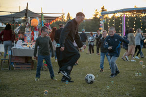 Ein junger Priester spielt Fußball mit Kindern auf einem Dorffest im Hintergrund Bäume und Mittagssonne. Bild aus dem Film