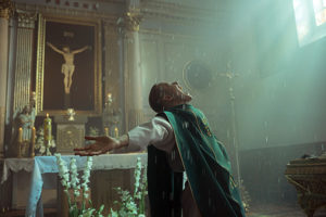 Ein junger Priester vor einem Alter seitlich stehend mit ausgestreckten Armen und von Sonnenlicht beleuchtet