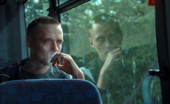 Ein junger Man sitzt im Bus und raucht, sein Gesicht spiegelt sich im Busfenster. Bild aus dem Film