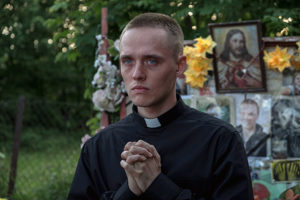 Ein junger Priester betet, im Hintergrund eine Tafel mit Blumen, Jesusbild und Trauerbilder. Bild aus dem Film