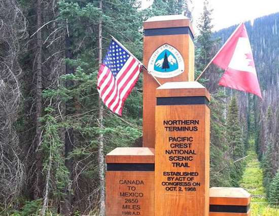 Holzmonument im Wald mit der Aufschrift: "Northern Terminus. Pacific Crest National Scenic Trail." US- und kanadische Flagge rechts auf das Monument aufgesteckt.