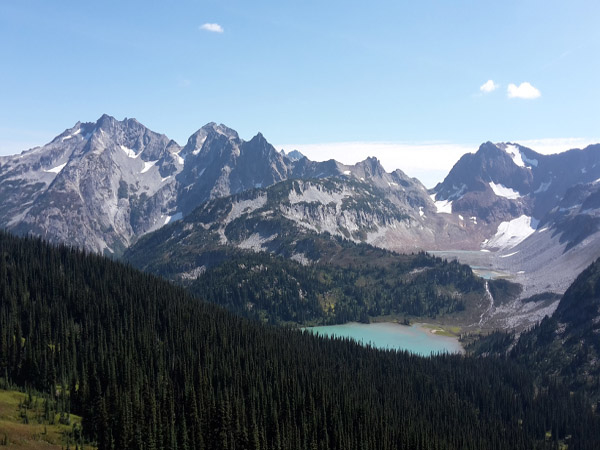 Bergpanorama mit Gletscherresten und einem türkisen See, im Vordergrund Nadelwald