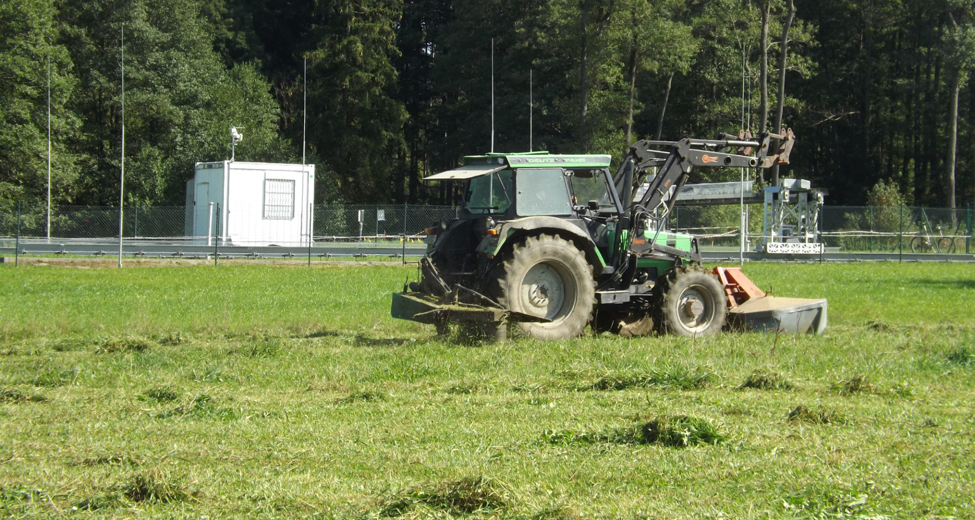 Ein Traktor mit Kreiselmäher fährt auf einer Wiese vor einer eingezäunten Meßstation für Klimaforschung im oberbayerischer Weiler Fendt