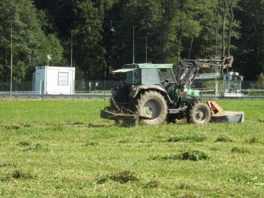 Ein Traktor mit Kreiselmäher fährt auf einer Wiese vor einer eingezäunten Meßstation für Klimaforschung im oberbayerischer Weiler Fendt